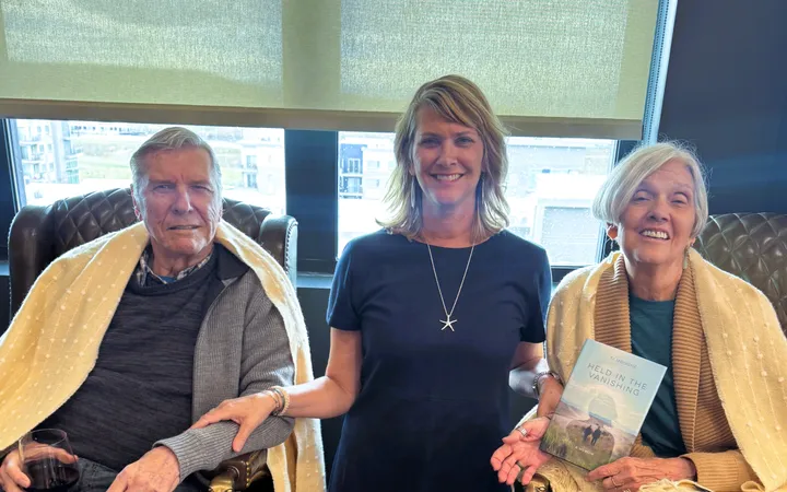 Ti Mougne standing between her father and mother, who are seated in leather armchairs wrapped in cream throws in a sunlit lounge; her mother holds a copy of Held in the Vanishing.