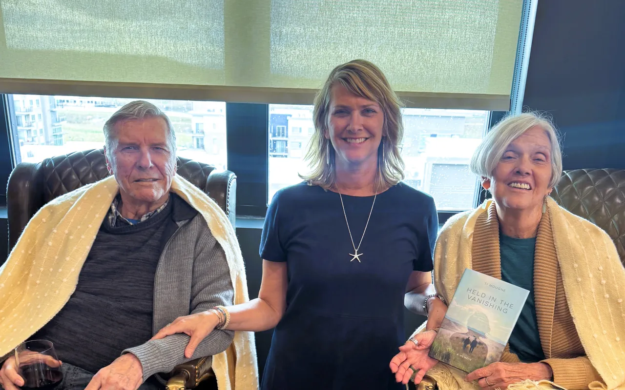 Ti Mougne standing between her father and mother, who are seated in leather armchairs wrapped in cream throws in a sunlit lounge; her mother holds a copy of Held in the Vanishing.