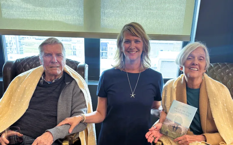 Ti Mougne standing between her father and mother, who are seated in leather armchairs wrapped in cream throws in a sunlit lounge; her mother holds a copy of Held in the Vanishing.
