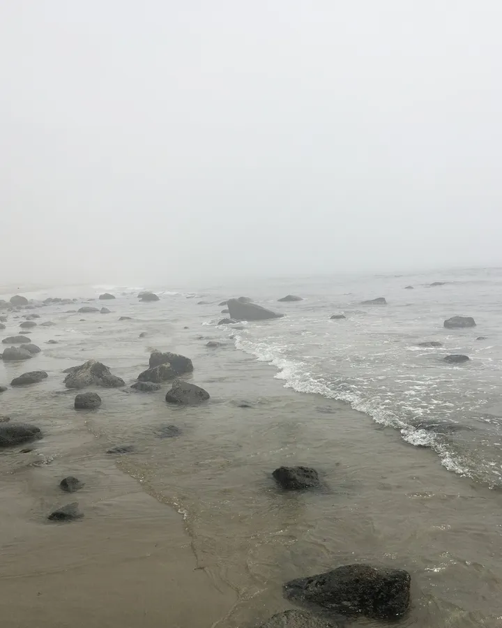 A rocky shoreline at the waterline, the sea and sky dissolving together into thick white fog.