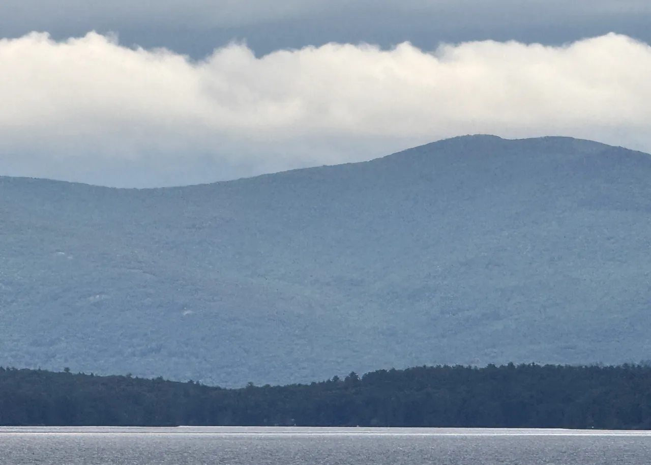 Forested mountains rising above the still, silvery water of Lake Winnipesaukee, their ridgeline softened by a low band of clouds.