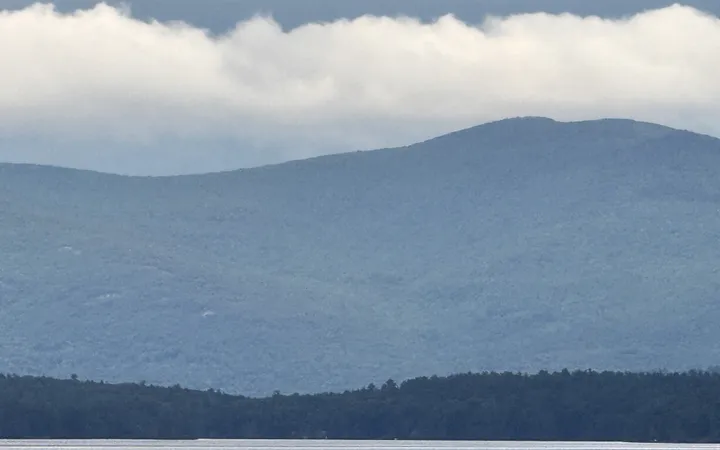 Forested mountains rising above the still, silvery water of Lake Winnipesaukee, their ridgeline softened by a low band of clouds.