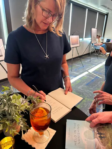 Ti Mougne, in glasses, signing a copy of Held in the Vanishing at her launch, an Aperol spritz and copies of the memoir on the table before her.