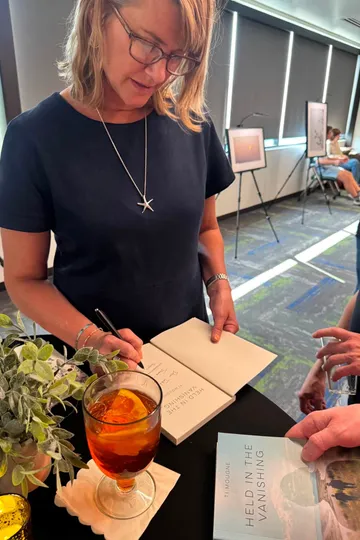 Ti Mougne, in glasses, signing a copy of Held in the Vanishing at her launch, an Aperol spritz and copies of the memoir on the table before her.