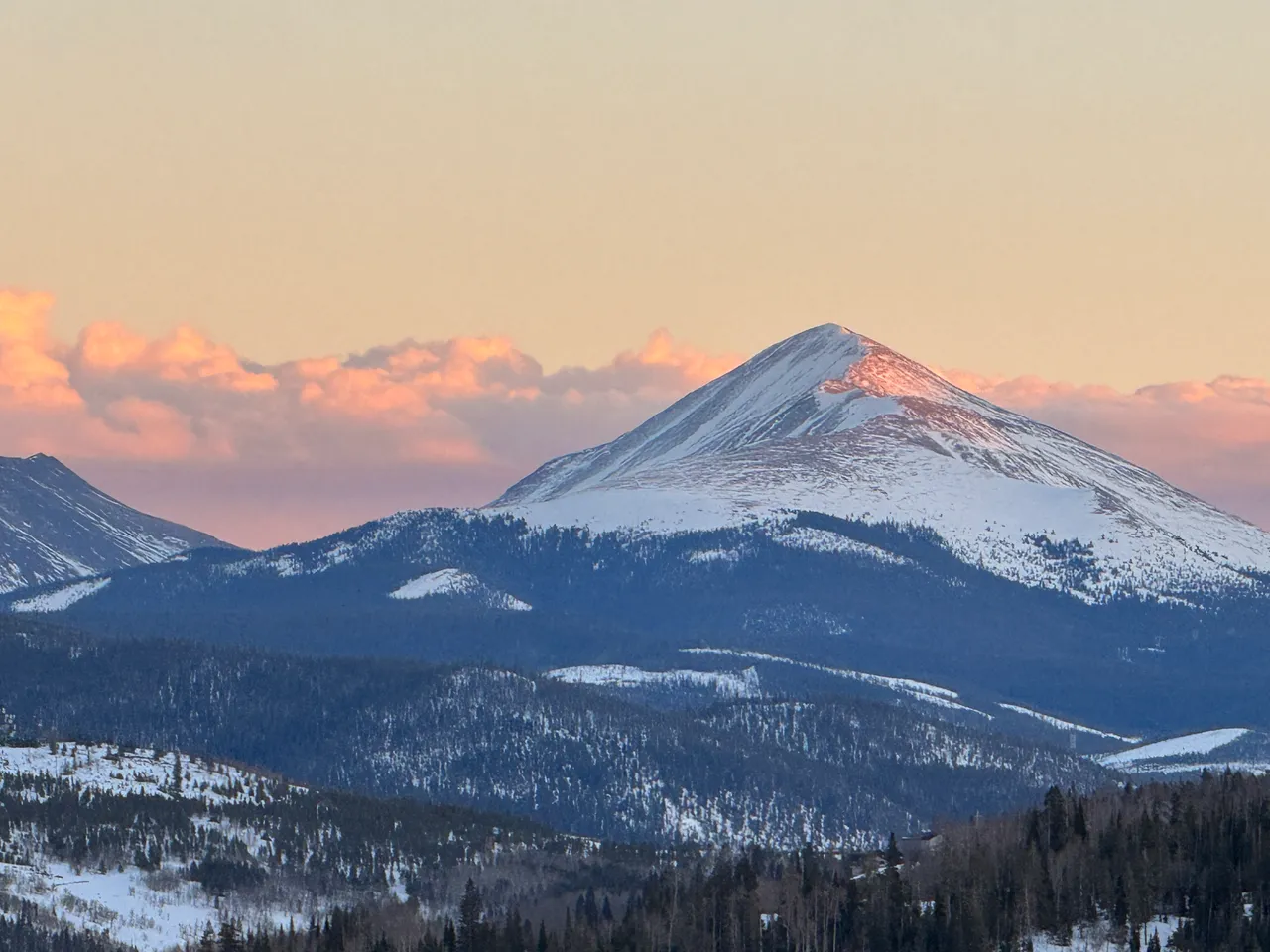 A snow-capped peak catching the last pink light of sunset above dark forested ridges, with rose-lit clouds along the horizon.