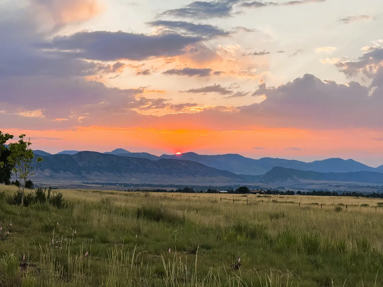 The sun slips behind a ridge of blue foothills at dusk, beneath clouds lit orange and violet, with a wide grassy meadow and a fence line in the foreground.