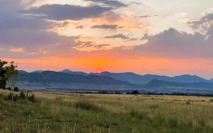 The sun slips behind a ridge of blue foothills at dusk, beneath clouds lit orange and violet, with a wide grassy meadow and a fence line in the foreground.