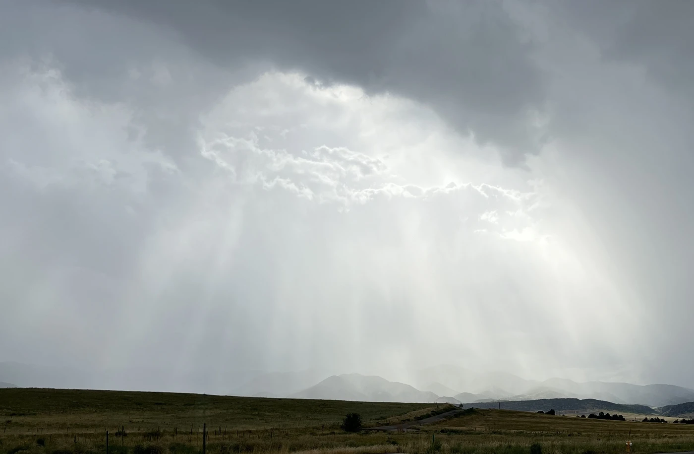 Sunlight breaking through a heavy grey sky in soft rays of rain over distant mountains and open prairie