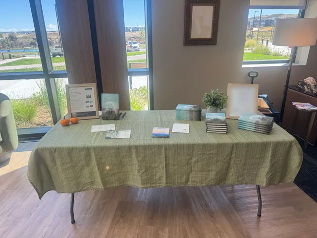 Ti Mougne's book table at The Reserve's Wellness Fair, draped in a sage-green cloth and set with stacks of Held in the Vanishing, framed signs, reflection cards, and two small pumpkins before tall sunlit windows.