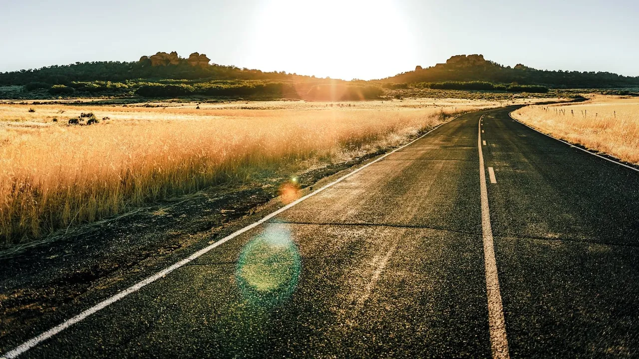 An empty road stretching through golden grassland toward sunlit hills at sunrise.